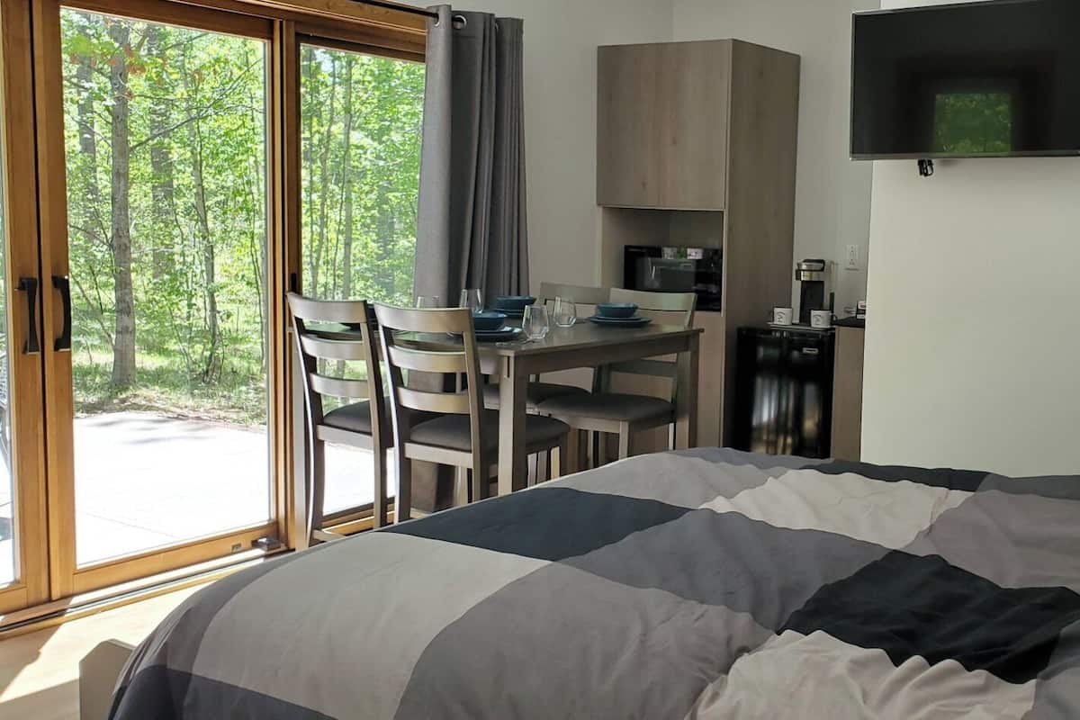 A bedroom with a bed, a desk, and a chair. There is a window with curtains, a flat-screen TV mounted on the wall, and a lamp on the side table.