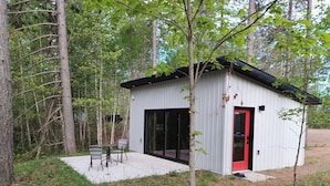 A small cabin with a red door, a patio with chairs, and a forested backdrop. - Expedition Inn (Calumet)