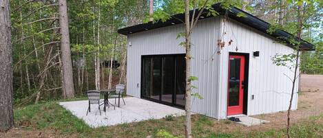 Eine kleine Holzhütte mit roter Tür, einer Terrasse mit Sitzgelegenheiten und einem Wald im Hintergrund.