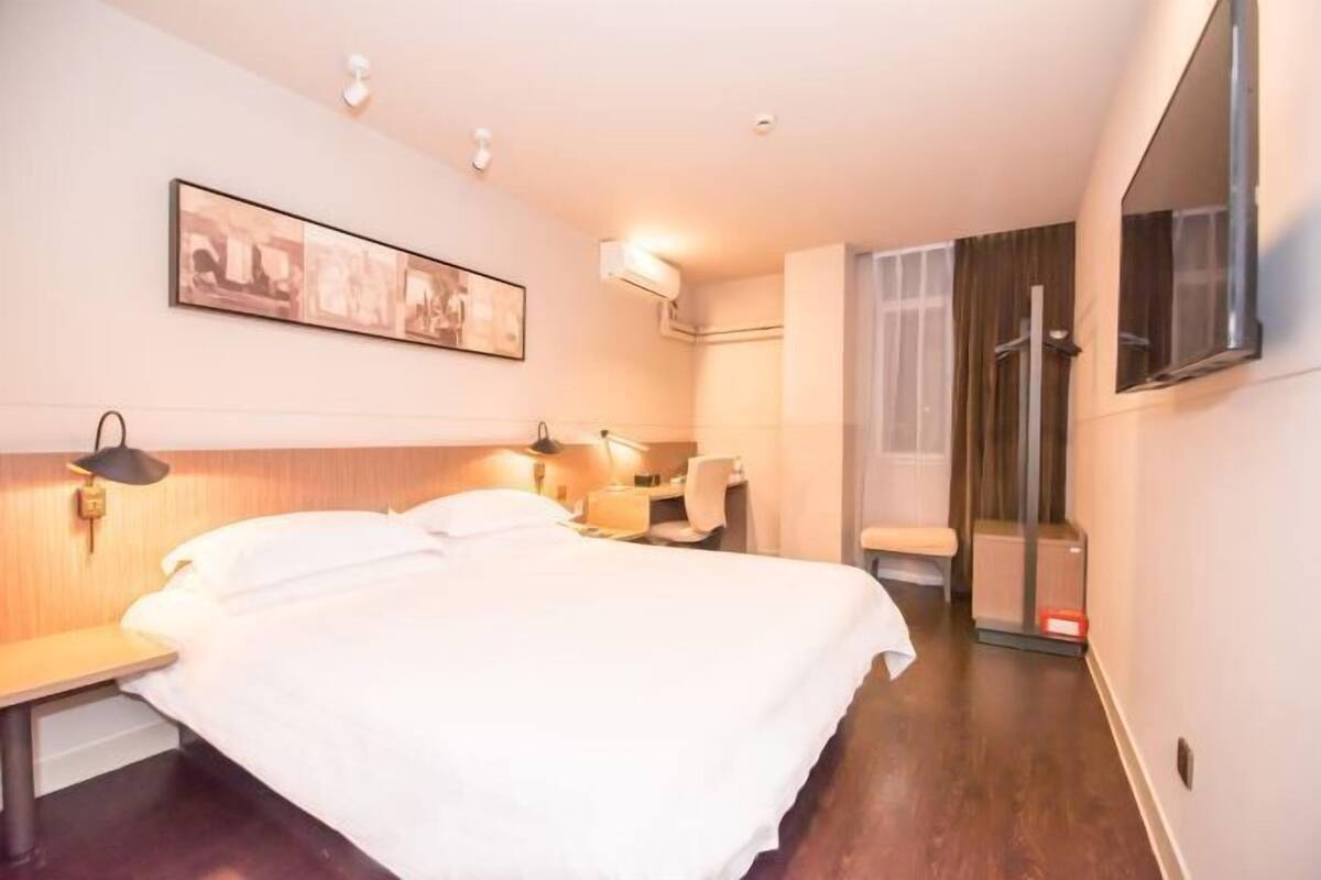 A hotel room with a bed, bedside table, lamp, wall-mounted artwork, and a shower area.