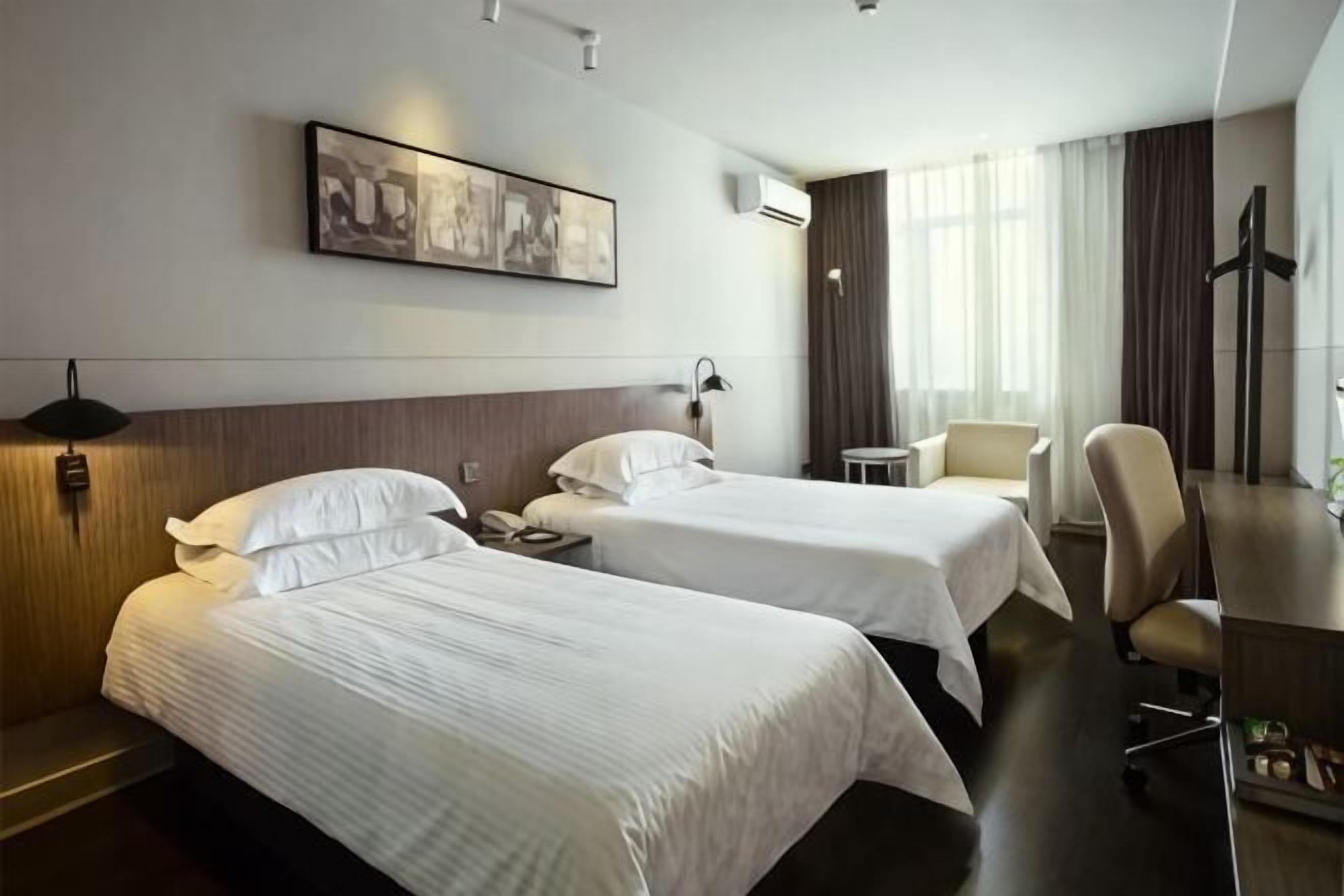 A hotel room with two beds, a desk, a chair, a small table, a wall-mounted artwork, and a cross on the wall.