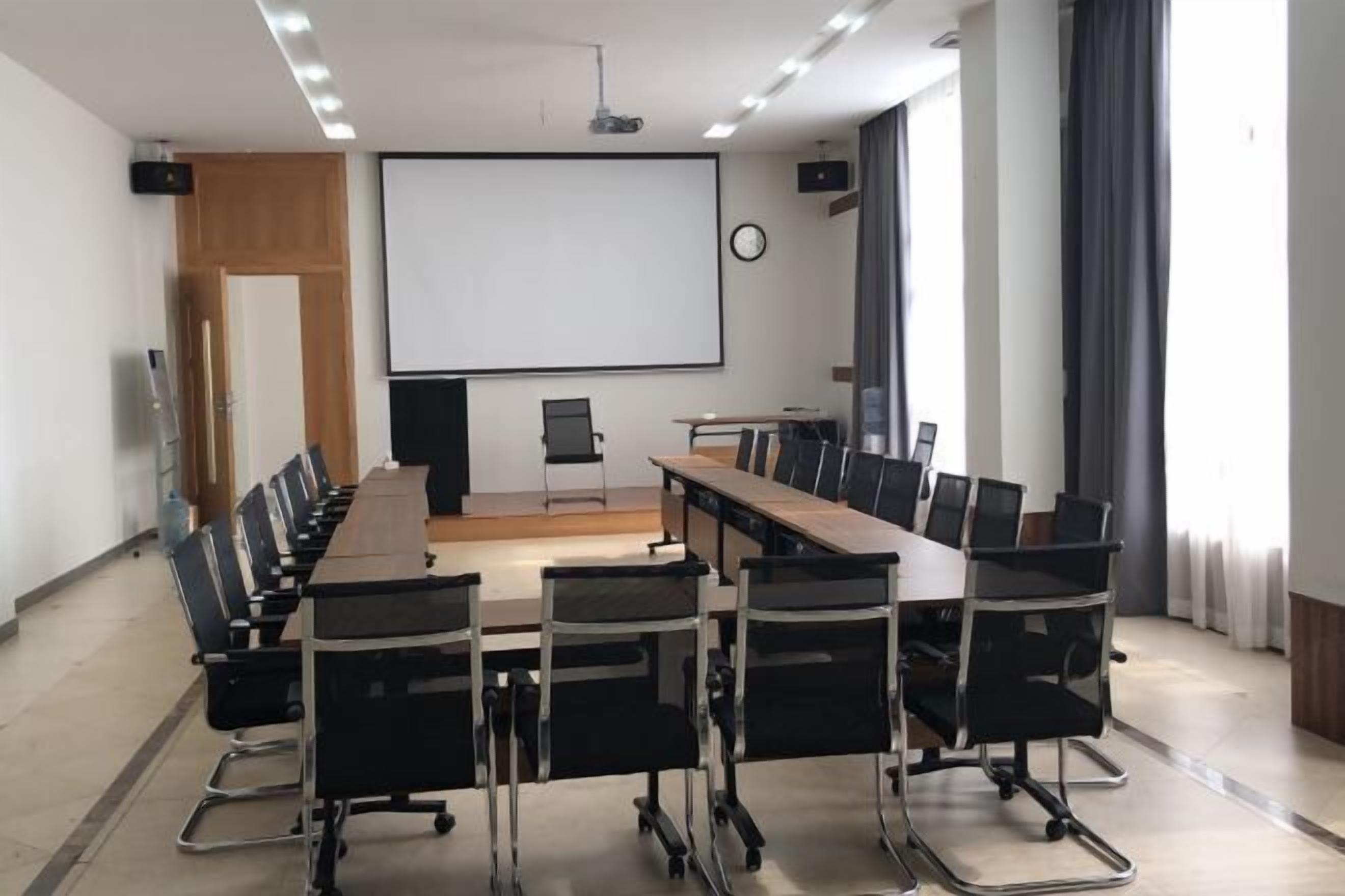 A conference room with a long table, chairs, a projector screen, and a clock on the wall.