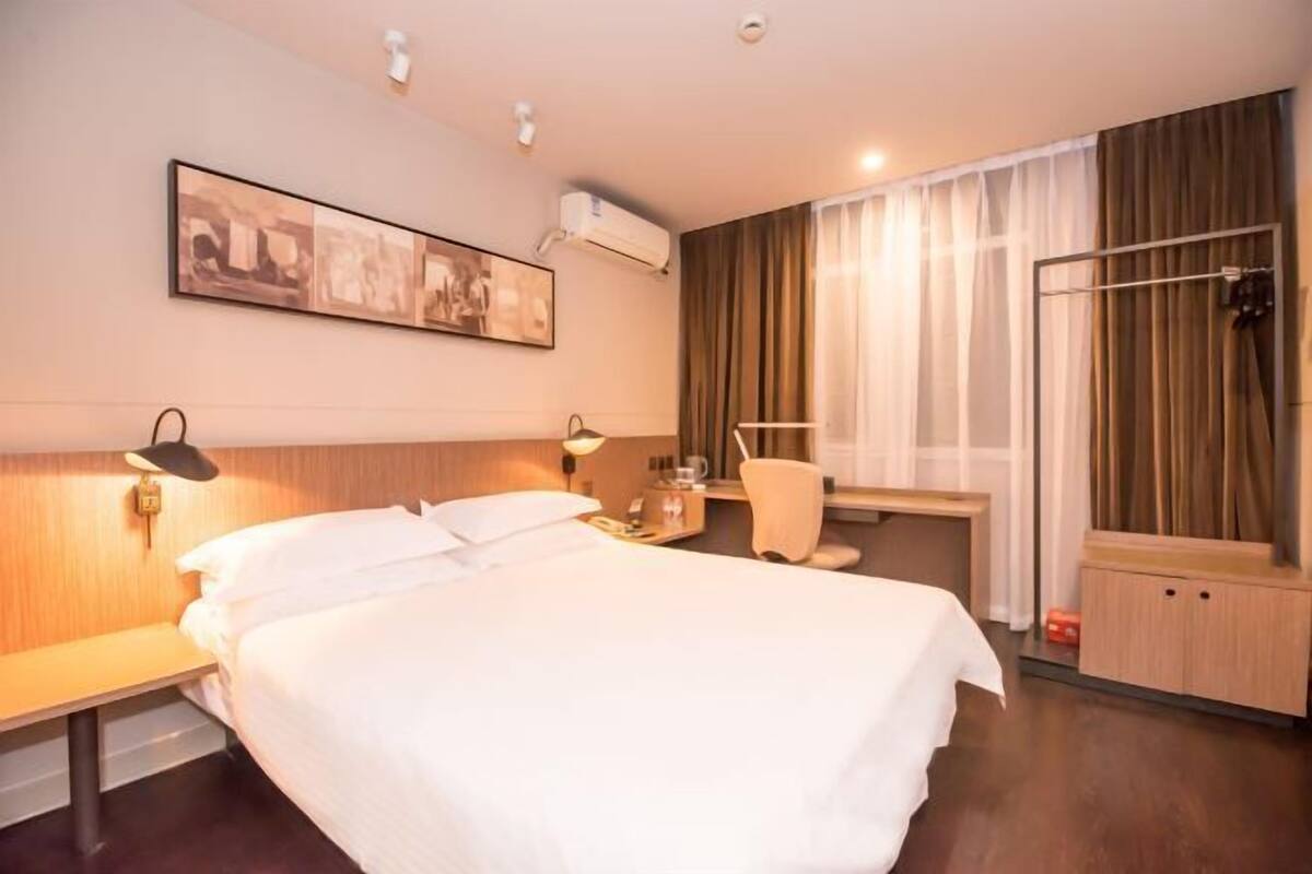 A hotel room with a bed, bedside table, lamp, air conditioning unit, and a shower area.