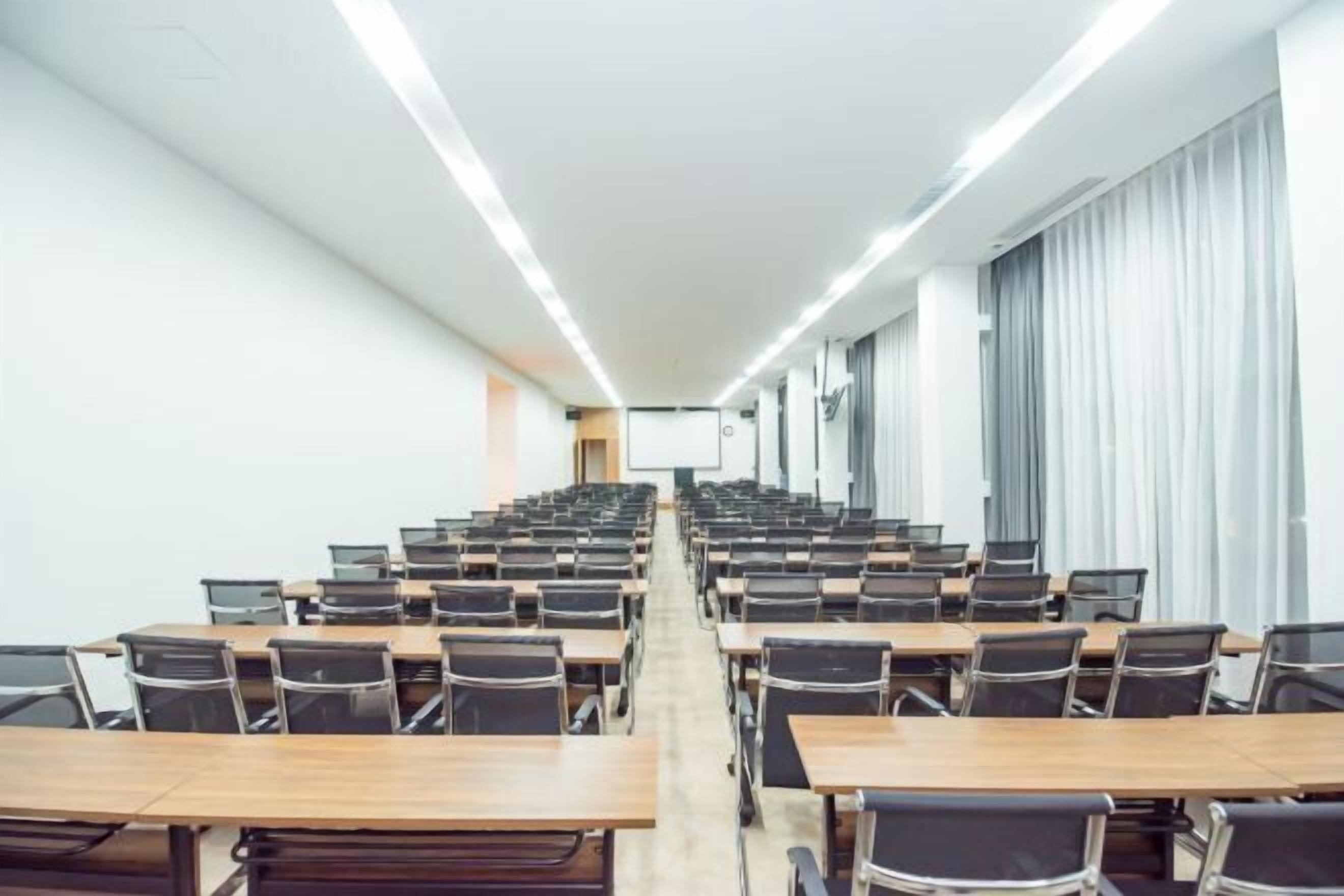 A conference room with rows of tables and chairs, a projector screen, and curtains on the windows.