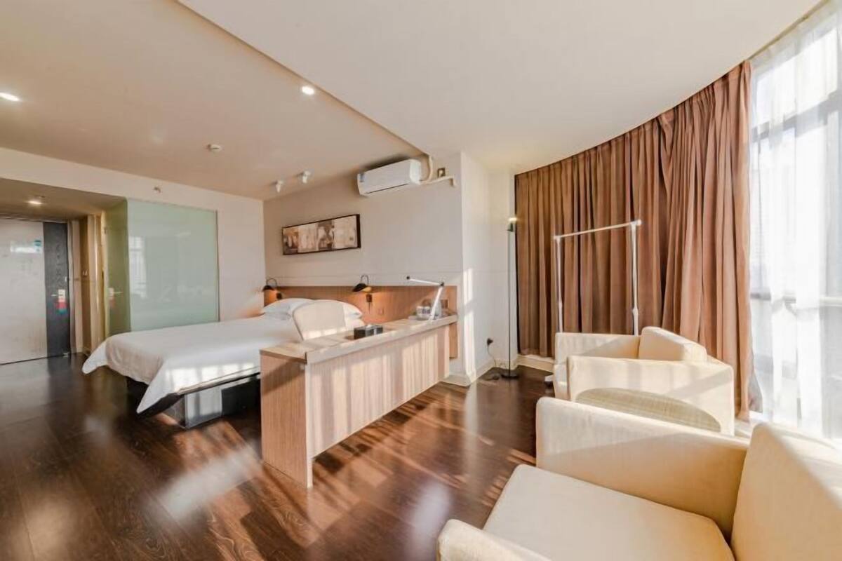 A hotel room with a bed, desk, chair, and large windows with curtains.
