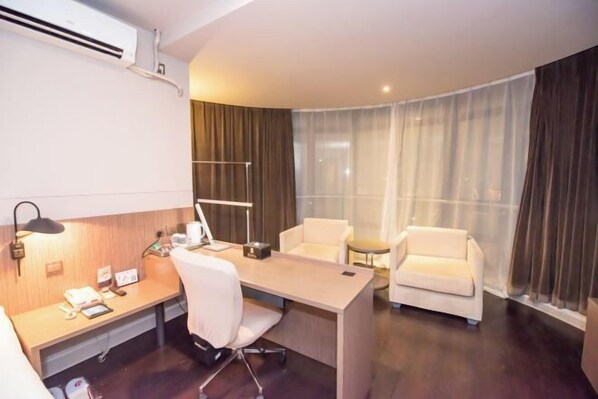 Quarto de hotel com escrivaninha, duas cadeiras, abajur, mesa pequena e janela com cortinas.