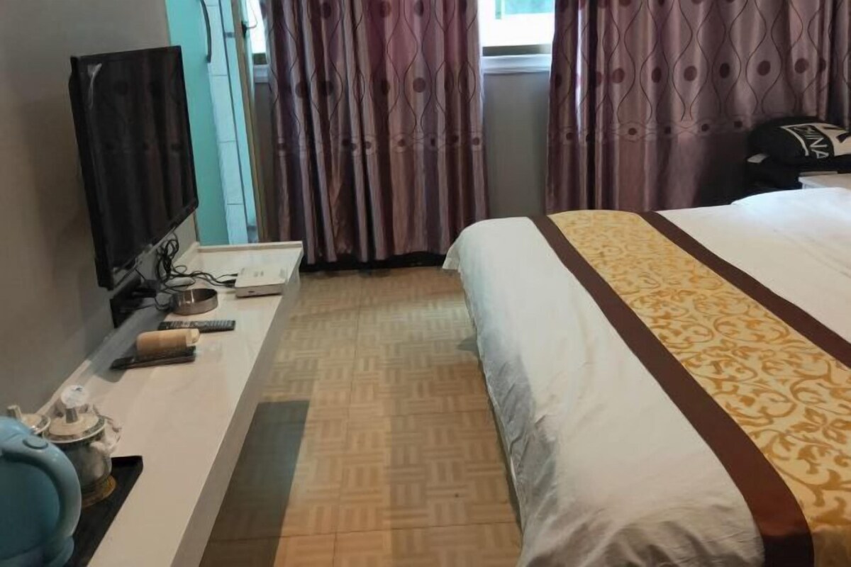 Ein Hotelzimmer mit einem Bett, einem Fernseher, einem Schreibtisch mit Computer und weiteren Gegenständen sowie gemusterten Vorhängen.