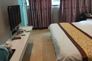 Ein Hotelzimmer mit einem Bett, einem Fernseher, einem Schreibtisch mit Computer und weiteren Gegenständen sowie gemusterten Vorhängen.