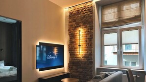 A modern living room with a brick accent wall, a flat-screen TV mounted on the wall, a grey sofa, a round coffee table, and a window with blinds. - Charm & Comfort Renovated Gem in Old Town 8 min walk to Krakow’s Main Square!  (Kraków)
