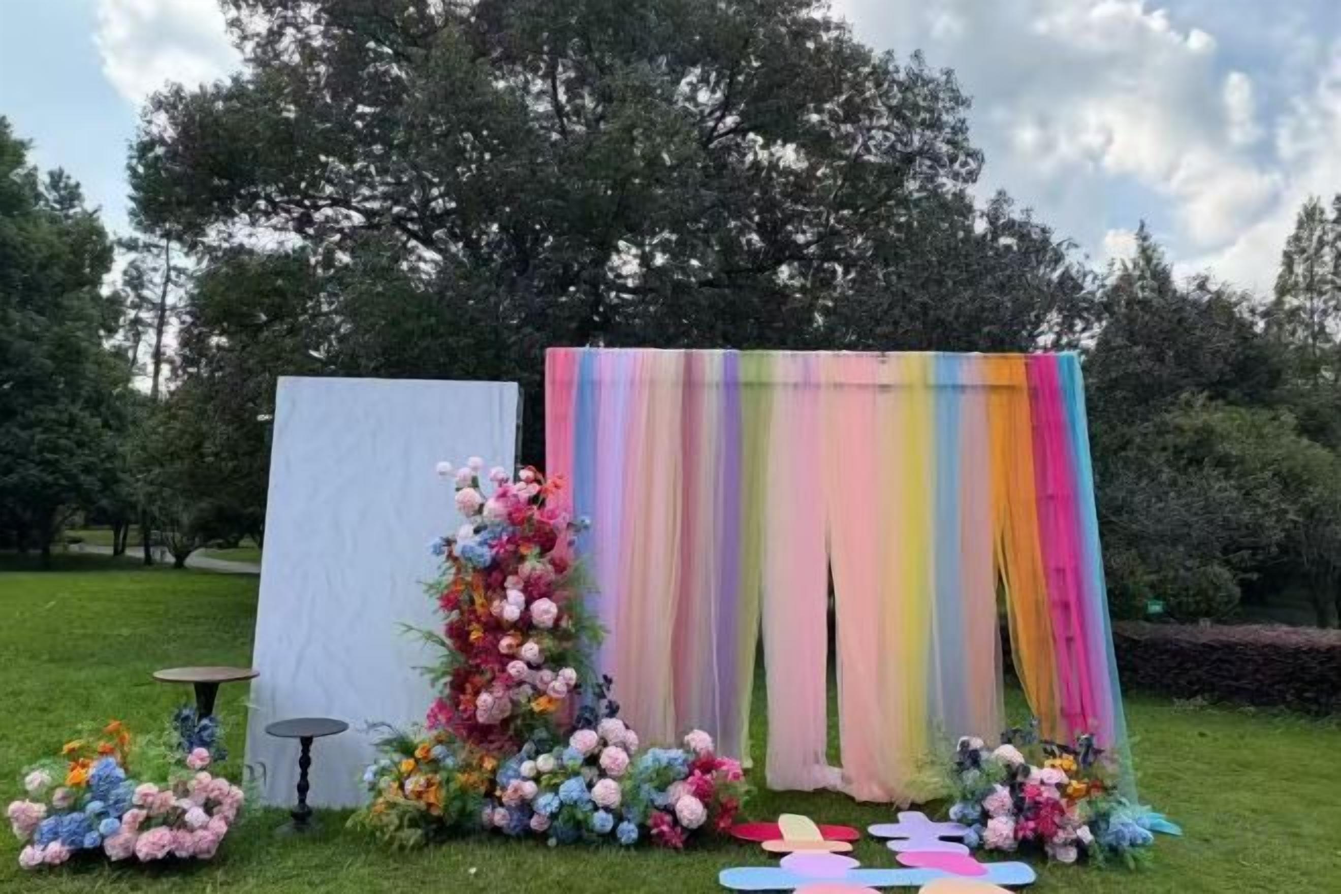 Eine Außenveranstaltungseinrichtung mit weißem Hintergrund und einem farbenfrohen Vorhang, umgeben von floralen Arrangements und einem kleinen Tisch.