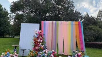 Eine Außenveranstaltungseinrichtung mit weißem Hintergrund und einem farbenfrohen Vorhang, umgeben von floralen Arrangements und einem kleinen Tisch.