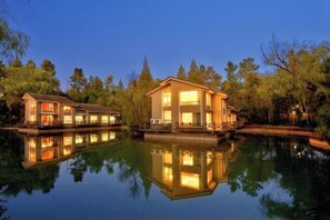 Une propriété située au bord de l’eau, comprenant deux bâtiments principaux, un bassin aux eaux calmes et des arbres environnants.