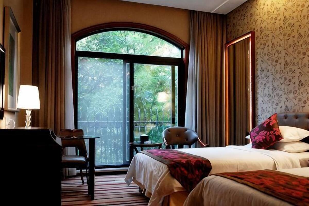 A hotel room with a bed, a desk, a chair, a balcony with a view of greenery, and a large window.
