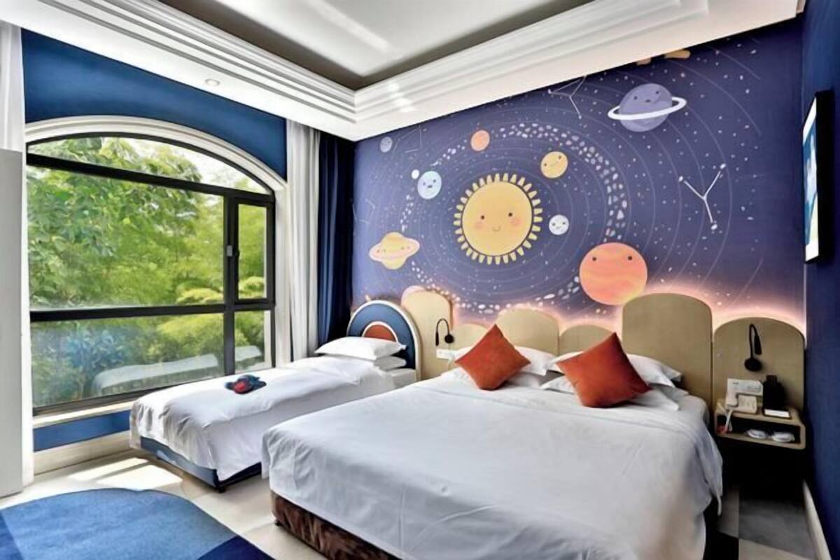 Un dormitori amb dos llits, una finestra gran i un mural al mur amb planetes.