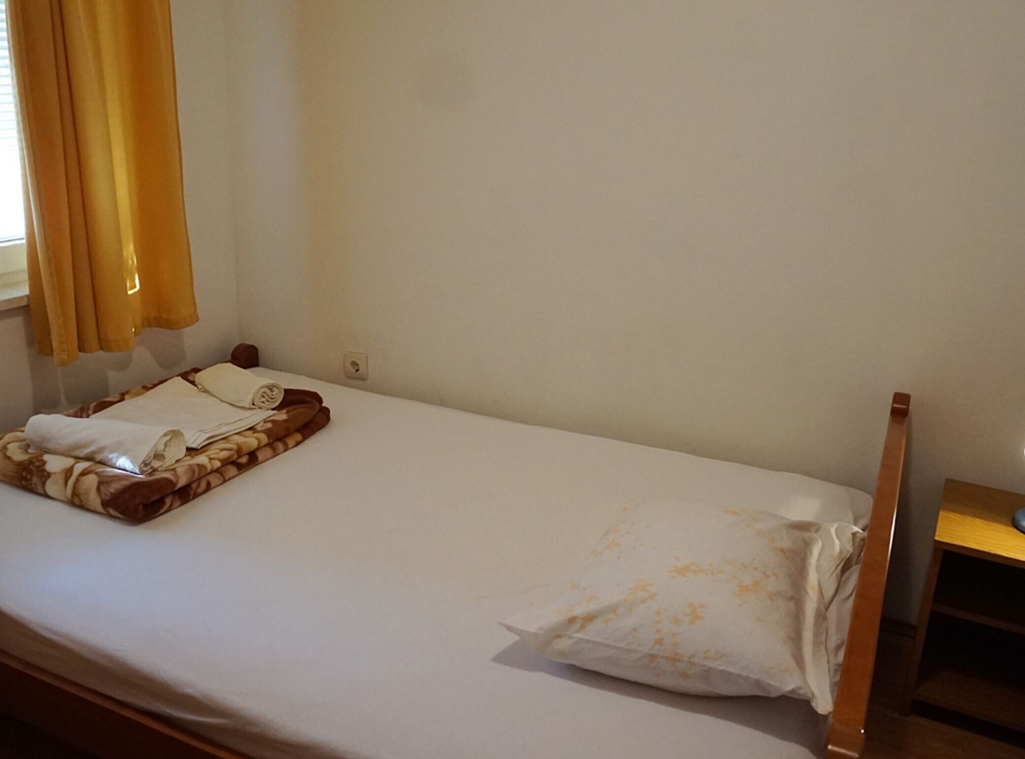 Een enkel bed met een wit laken, een bruine deken en een kussen. Rechts staat een houten nachtkastje.