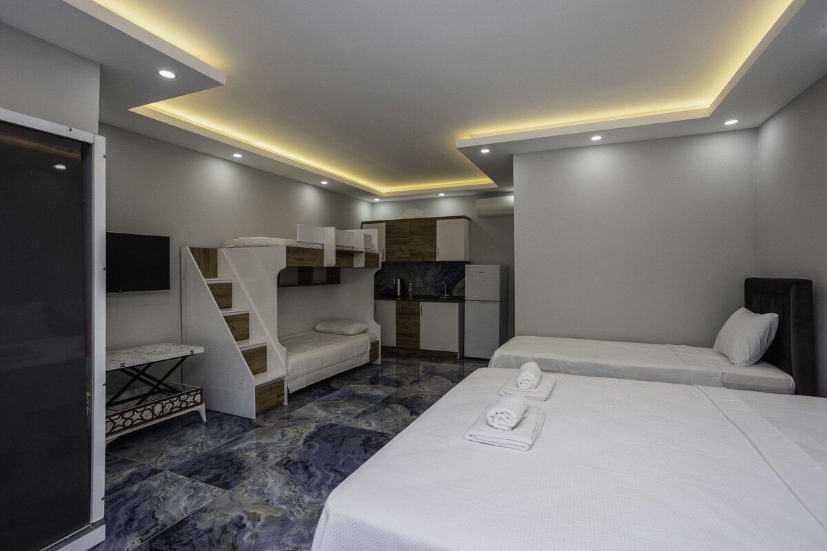 Quarto de hotel moderno com uma cama grande, uma cama menor, área de estar, cozinha compacta e televisão.