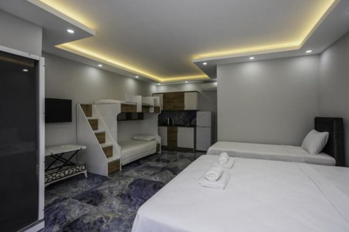 A modern hotel room with a bed, a sofa, a TV, and a small kitchen area.