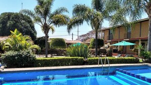 A swimming pool with steps leading into it, surrounded by palm trees and a building with a tiled roof. - Casa Akbal (Malinalco)