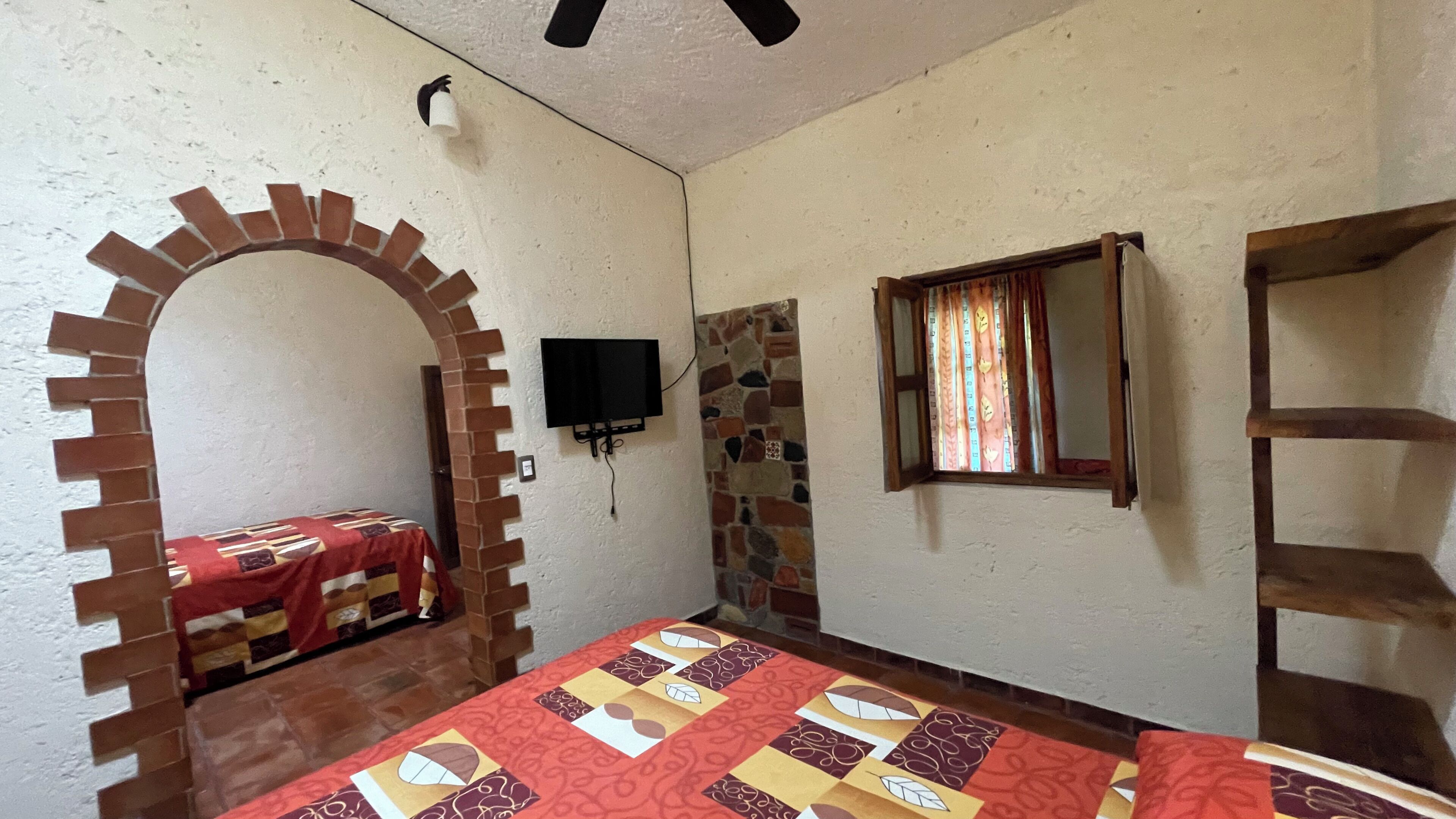 A room with a brick archway, a bed with a patterned bedspread, a television mounted on the wall, and a window with curtains.
