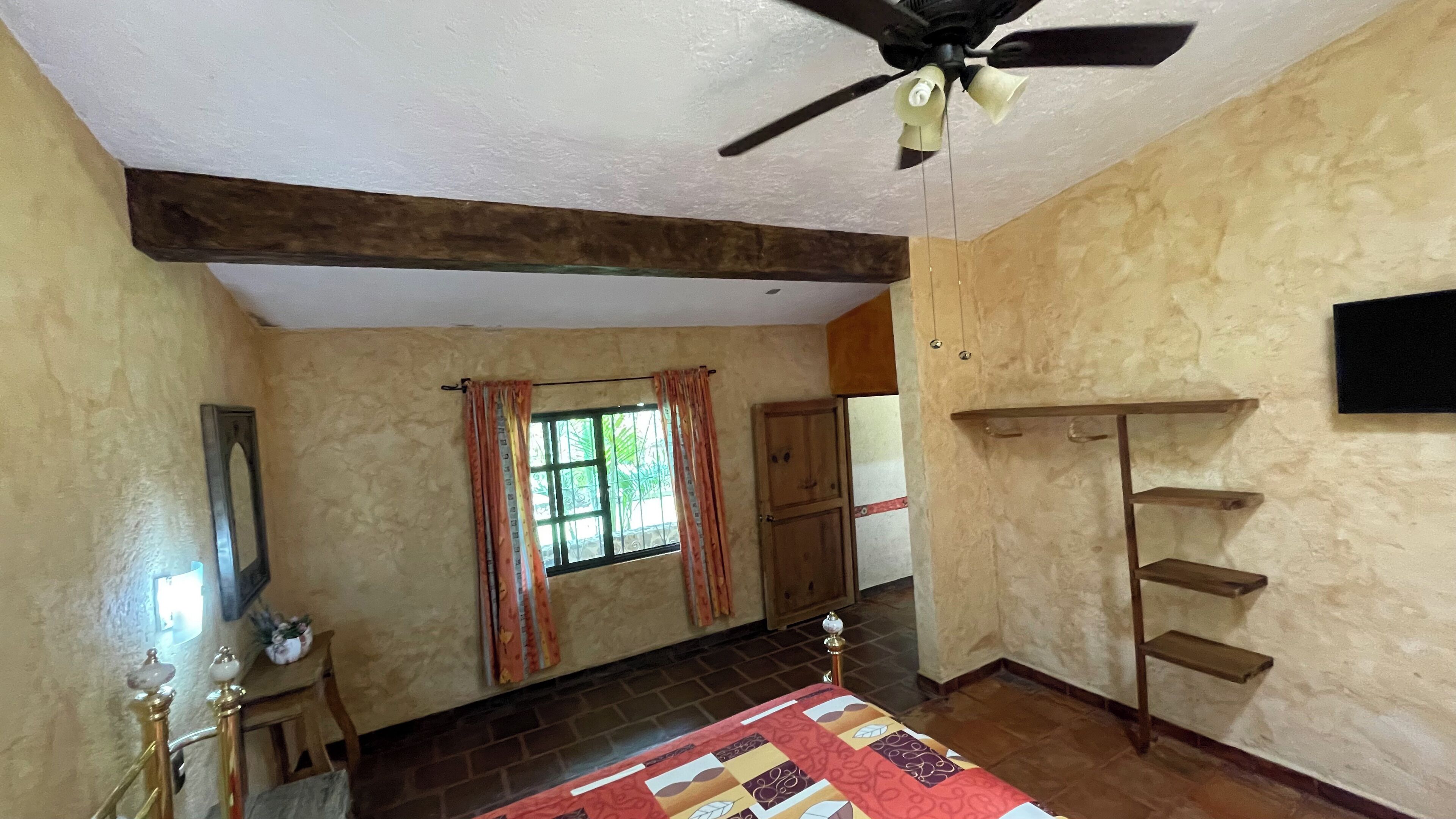 Une pièce avec un ventilateur de plafond, une fenêtre avec des rideaux, une porte en bois, une étagère et un lit recouvert d’une couvre-lit à motifs.