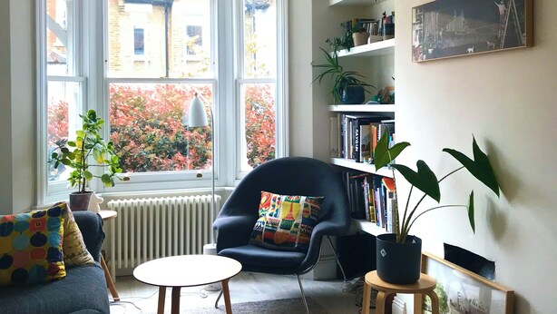 A living room with a large window, a black armchair, a small round table, and a bookshelf. - Cosy Victorian house in south London with a sunny private garden. (LONDON)