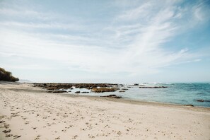 A beach with footprints leading to the water, rocky outcrops, and a clear sky.