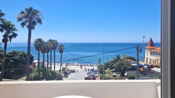 A balcony with a white table and chairs, overlooking a beach with palm trees and a clear blue sky.