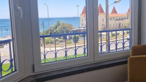 A room with large windows overlooking a coastal area, featuring a view of the sea and buildings with red roofs. - NEW! ESTORIL SEAFRONT 1BEDROOM APARTMENT (Estoril)