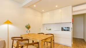 A compact kitchen with white cabinets, a wooden dining table, and chairs. There is a small plant on the table and a floor lamp beside it. - NEW! ESTORIL SEAFRONT 1BEDROOM APARTMENT (Estoril)