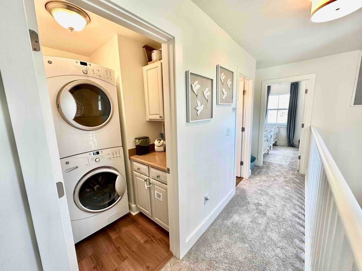 A laundry room with two stacked front-loading washers and dryers, a countertop with a basket, and a door leading to a hallway with a radiator and a window.