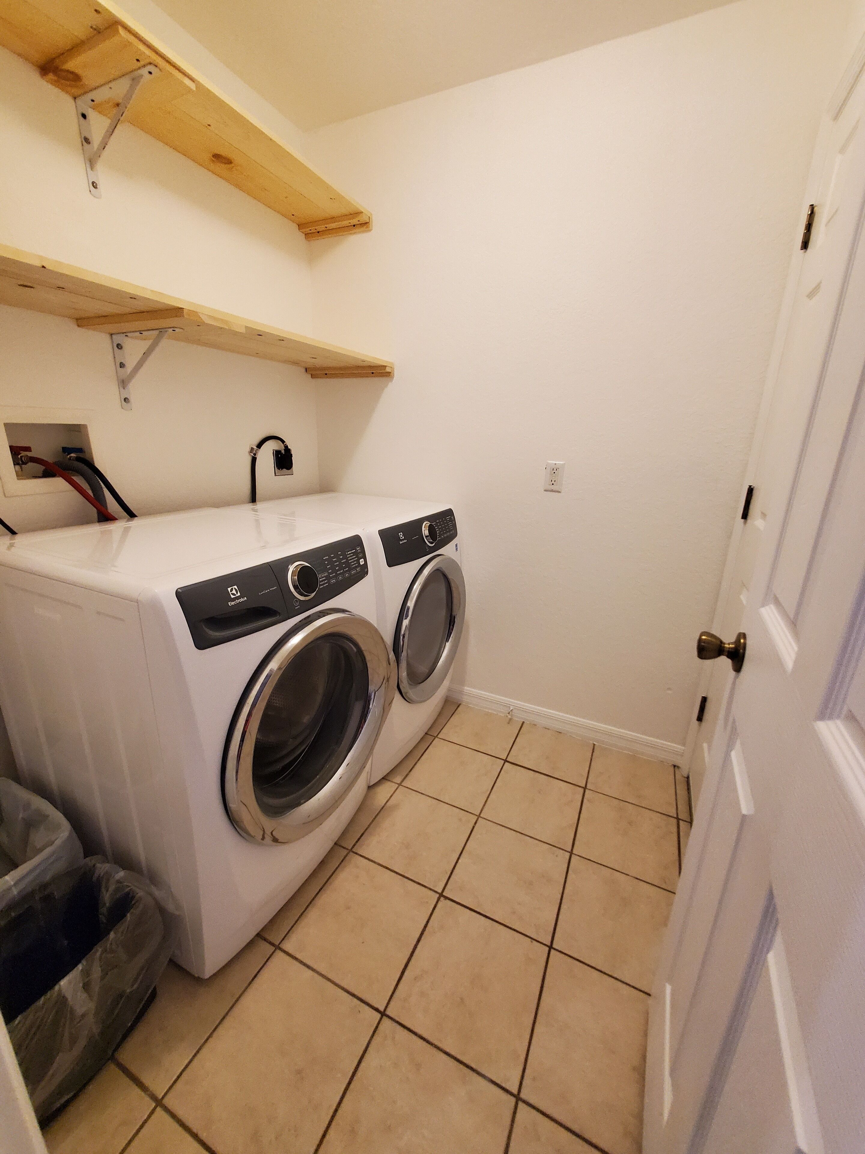 A small laundry room with two front-loading washing machines, a wooden shelf above, and a tiled floor.