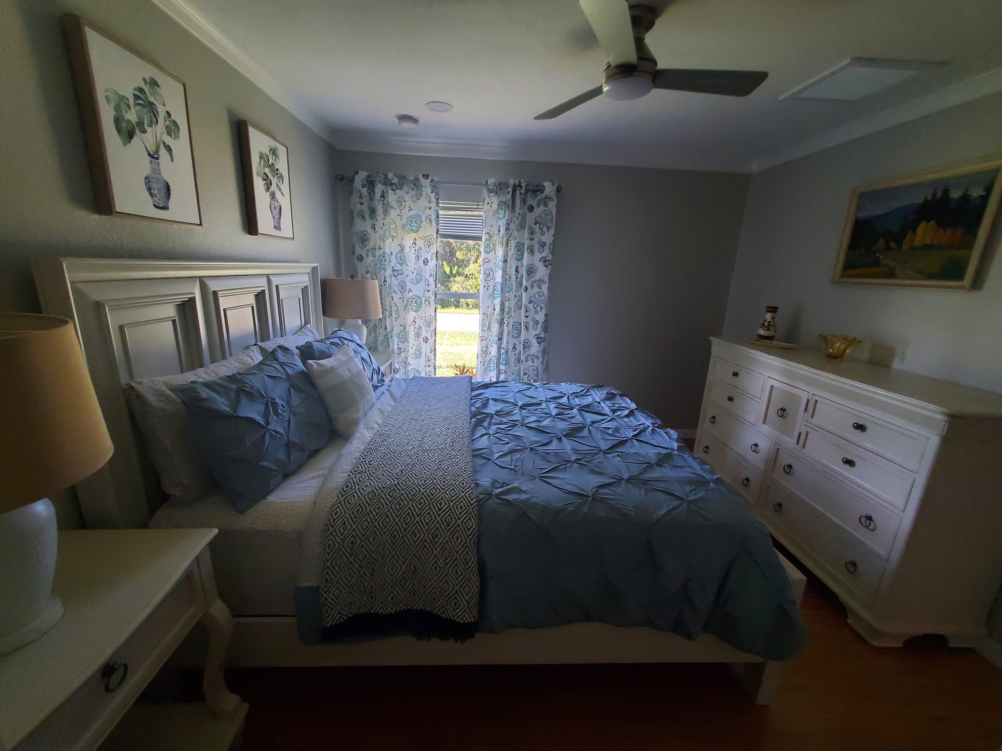 Un dormitorio con cama, cómoda, ventilador de techo y ventana con cortinas.