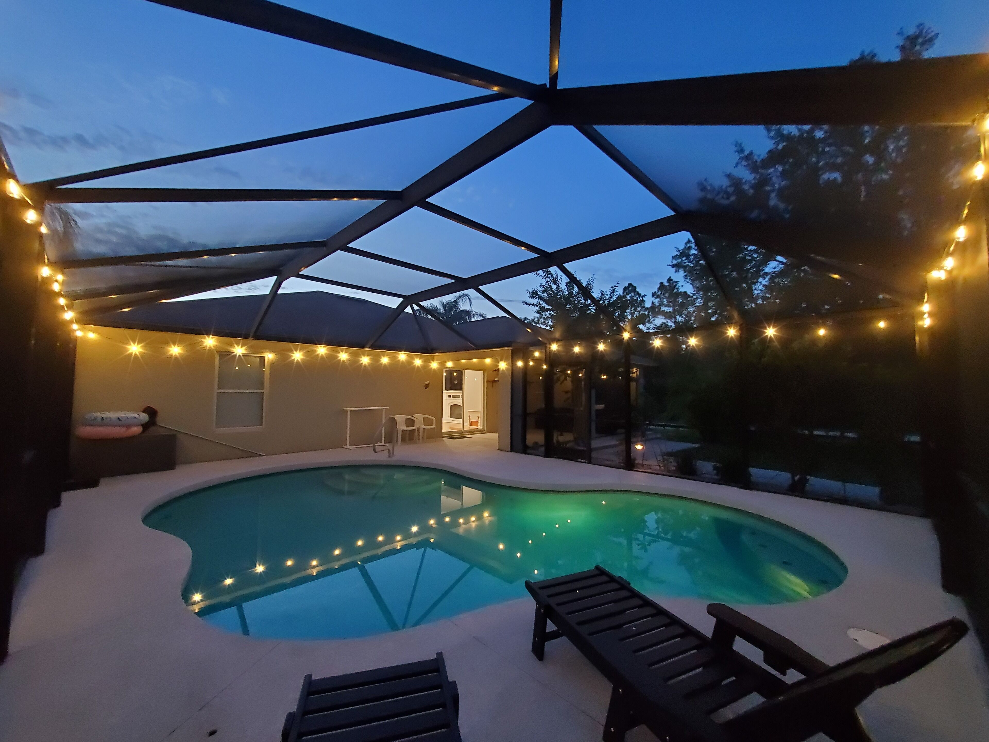 Una piscina con techo de vidrio, rodeada de luces colgantes, y un área de patio con sillones de descanso.
