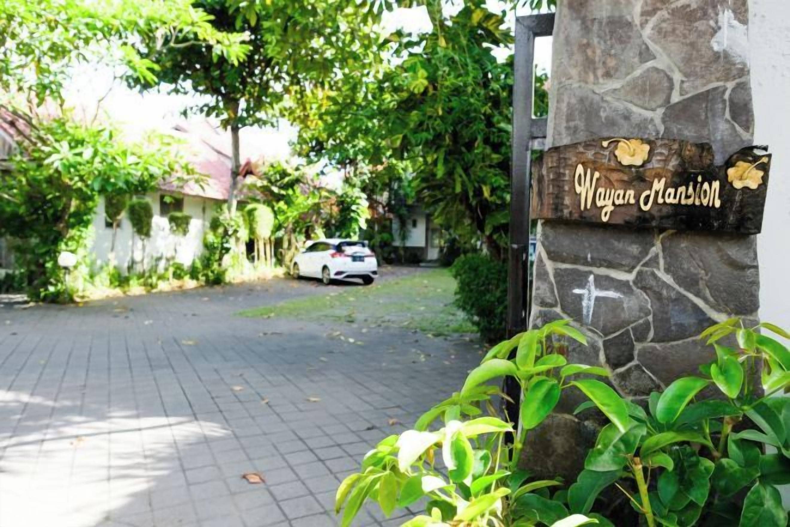 A paved driveway leading to a gated entrance with a stone wall and a sign reading 'Wayan Mansion.'