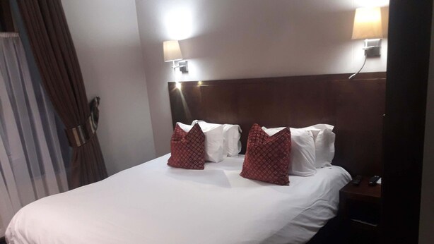 A hotel room with a large bed, two bedside lamps, a nightstand with a remote control, and a curtain on the left. - Potchefstroom Guest House with WiFi (Potchefstroom)