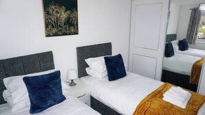 A hotel room with two beds, white bedding, dark blue pillows, a wooden throw blanket, a nightstand with a lamp, and a framed picture on the wall. - Biz & Fam Sleeps 7 Wigan w/ Parking, Self-check-in (Wigan)