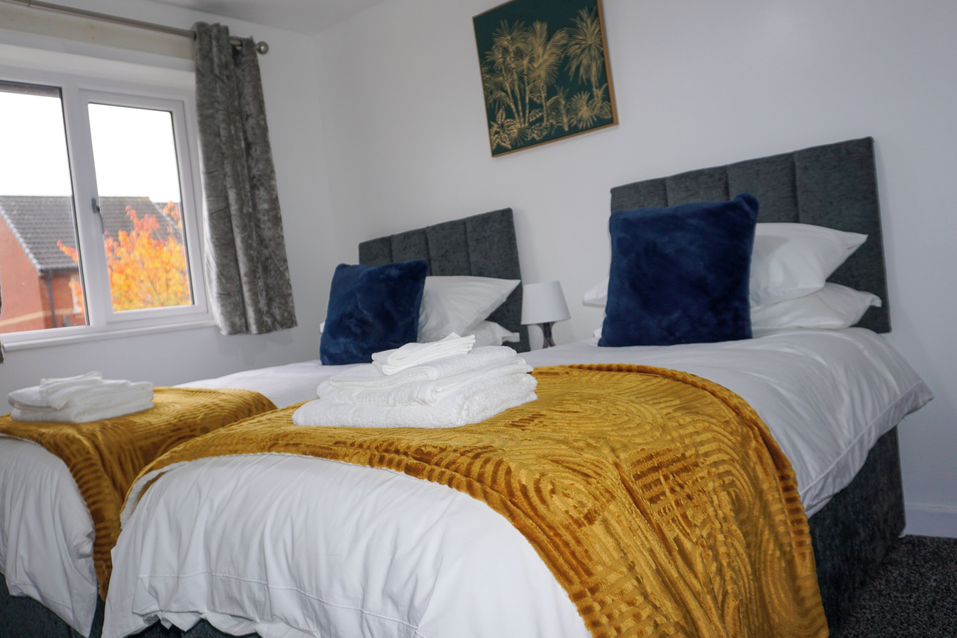 A hotel room with two beds, a window with curtains, a painting on the wall, and a view of trees outside.