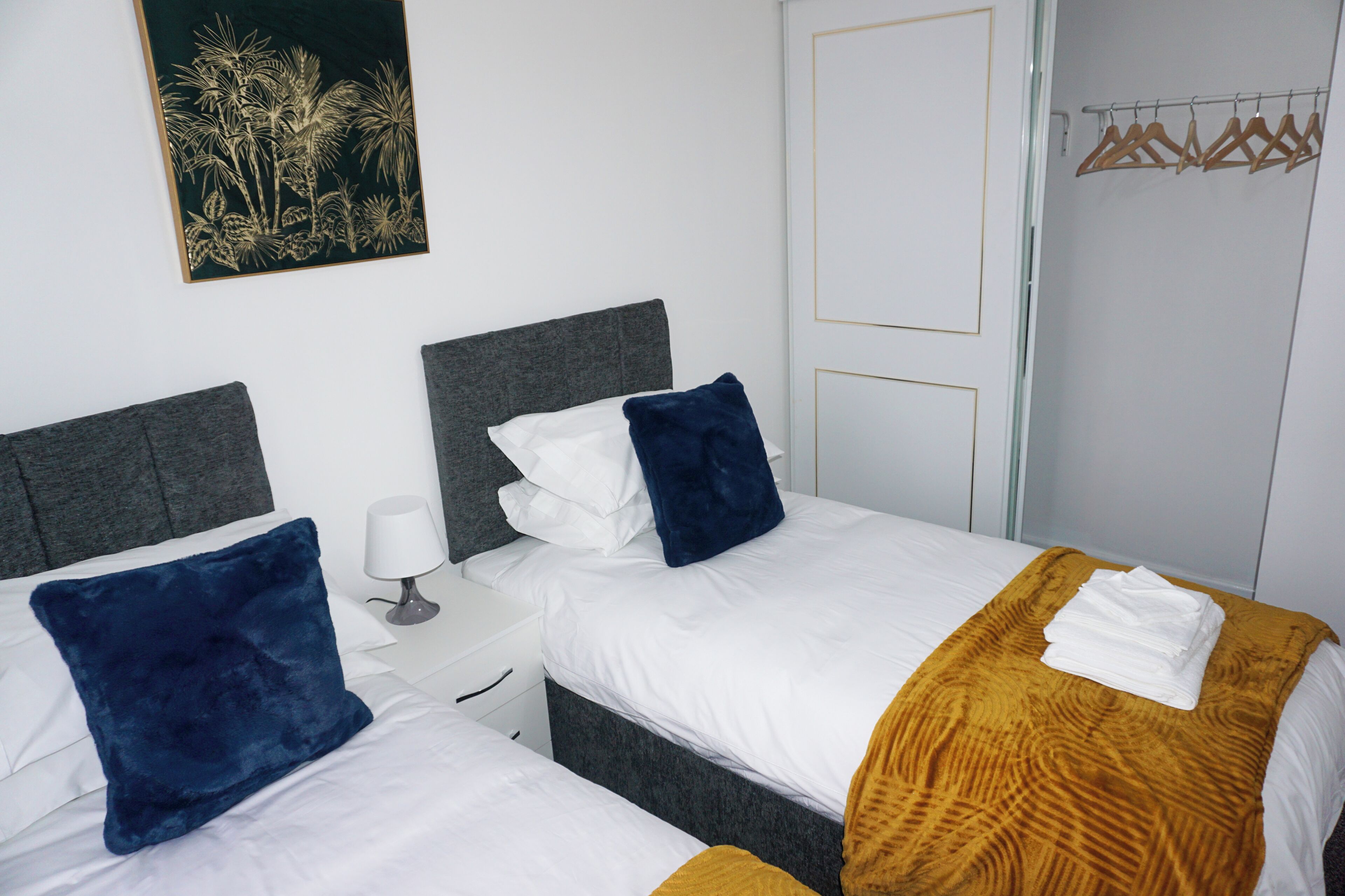 Une chambre d’hôtel avec deux lits, du linge de lit blanc, des oreillers bleu foncé, une tête de lit en bois, une table de chevet blanche, une lampe de chevet, un tableau accroché au mur et une armoire avec des cintres en bois.