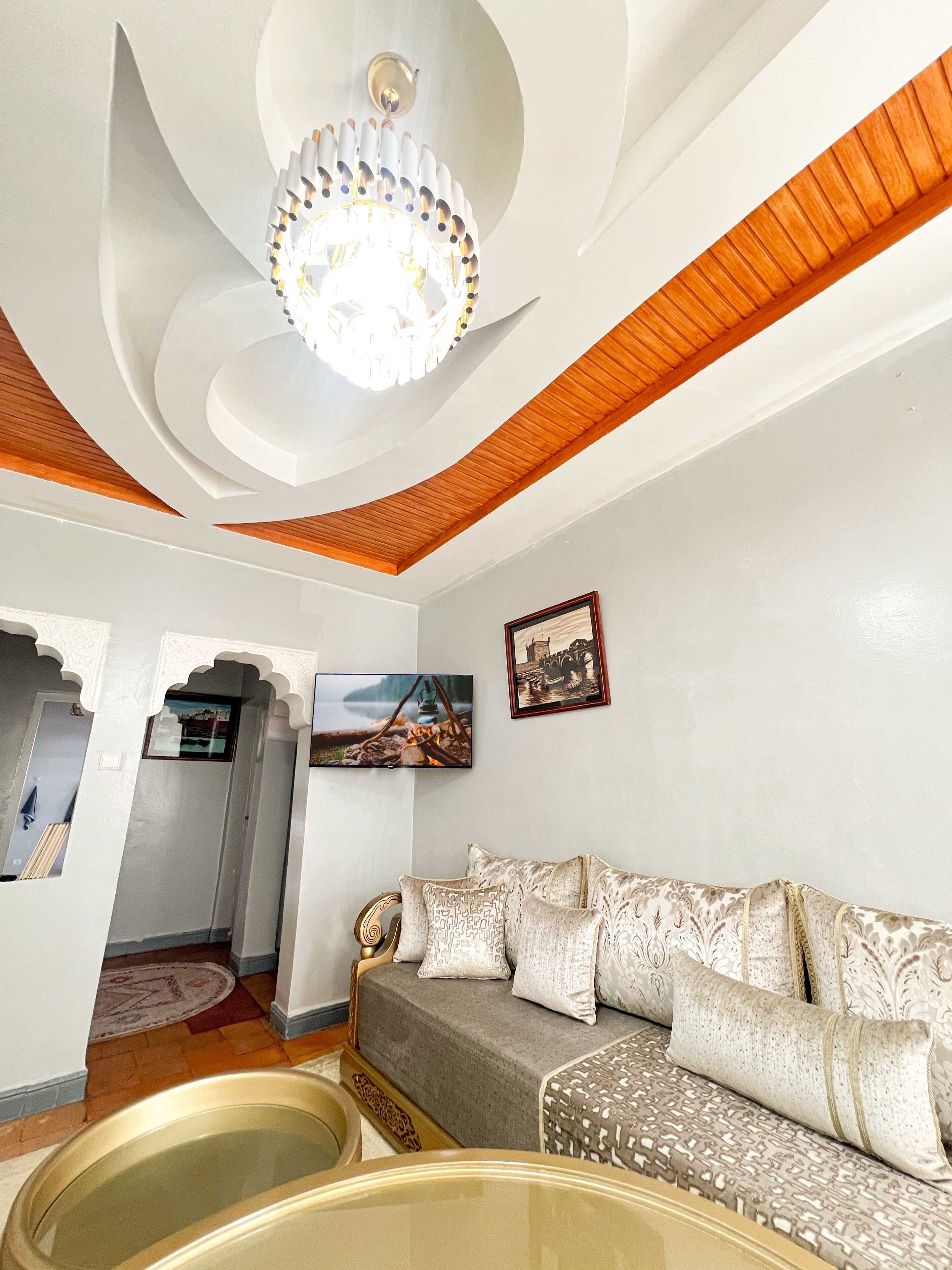 A living room with a modern chandelier, a sofa with patterned cushions, and a television mounted on the wall.