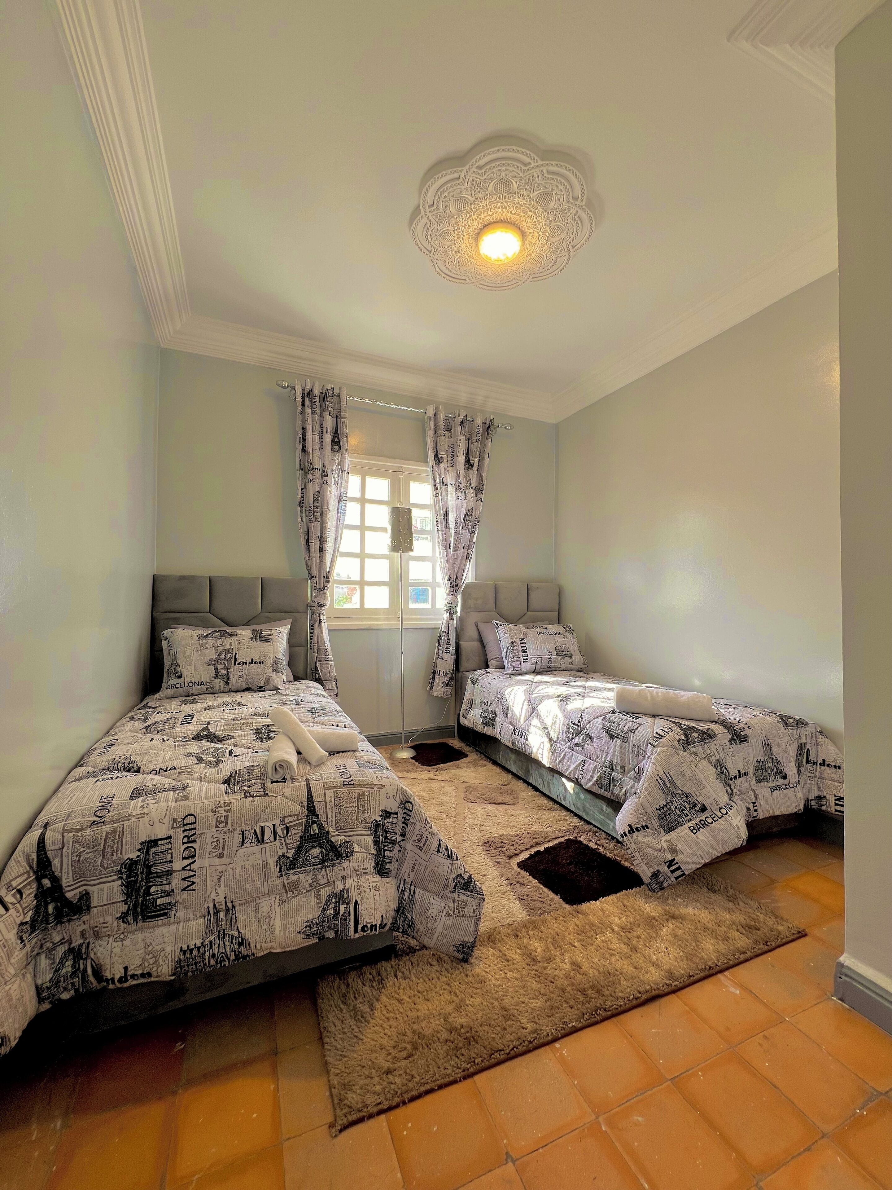 A bedroom with two beds, a decorative ceiling light, and a window with curtains.