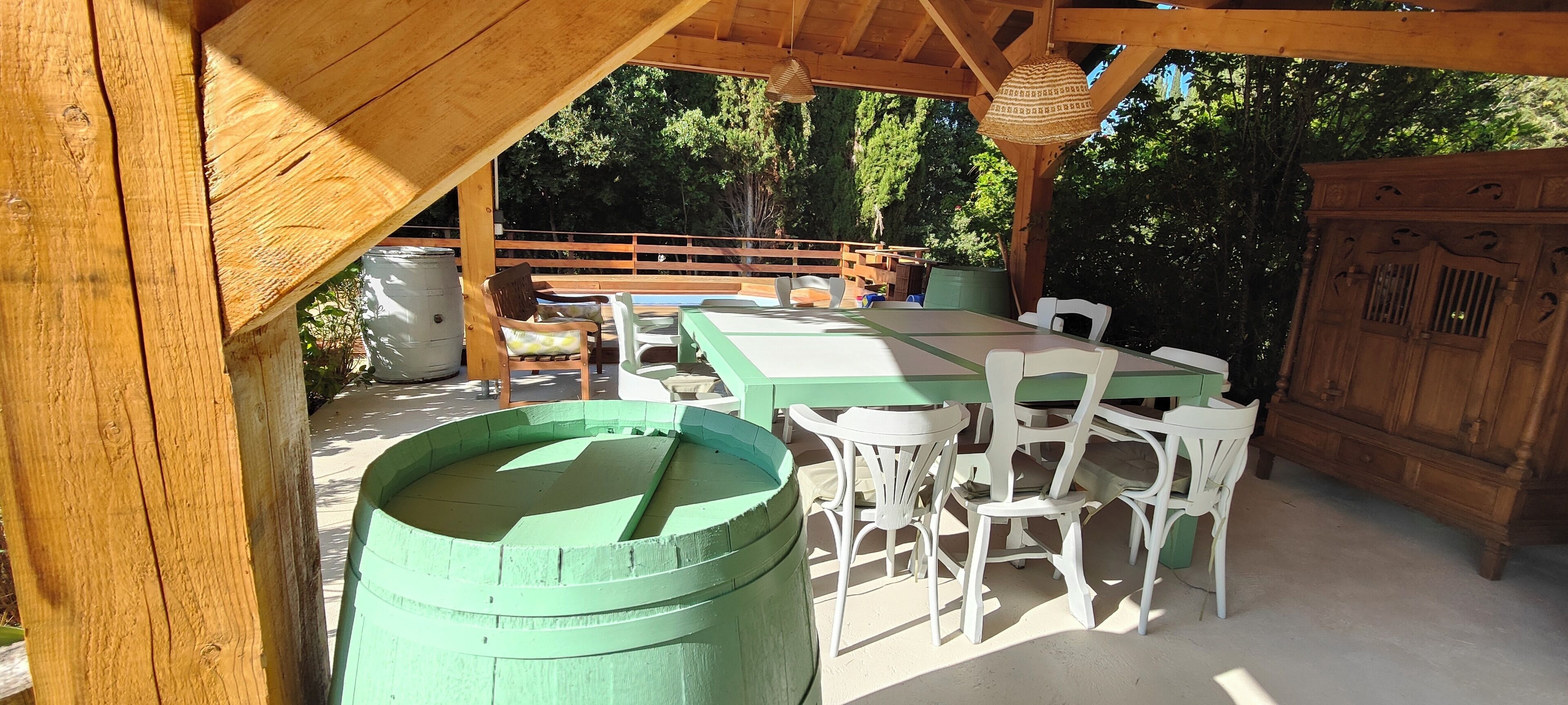 Une terrasse extérieure couverte, avec une table en bois et des chaises blanches, un tonneau vert et un meuble en bois.