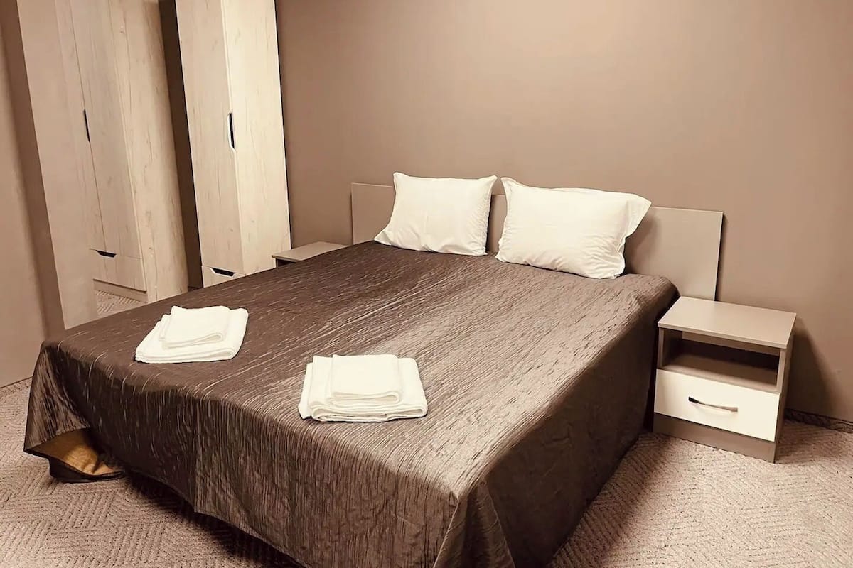 A neatly made bed with a dark brown bedspread and two pillows, a white nightstand with a drawer, and a tall white wardrobe.