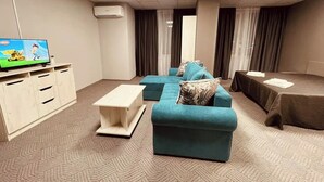 Habitación de hotel con sofá turquesa, una mesita blanca, televisor sobre un mueble de madera, cama con cubrecama oscuro y un ventanal con cortinas.