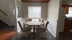 Un comedor con una mesa redonda y seis sillas, una ventana con persianas, y una cocina al fondo.