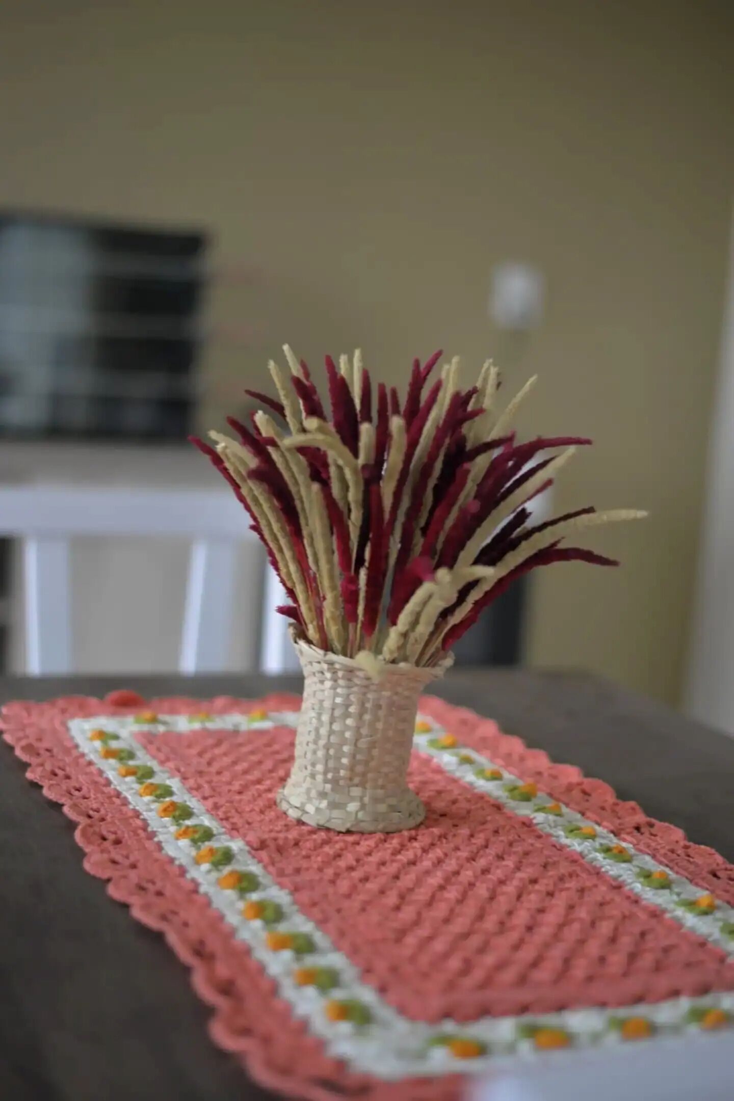 A table with a crocheted placemat and a woven vase with dried plants.