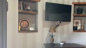 Uma sala de estar com televisão de tela plana na parede, prateleiras de madeira com objetos decorativos, uma luminária de vime sobre um móvel e uma poltrona.