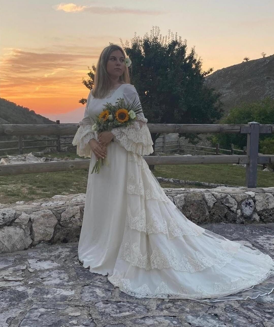 Une personne vêtue d’une robe blanche à longue traîne, tenant un bouquet de fleurs, se tient devant un mur de pierre, avec une clôture en bois et des arbres en arrière-plan.