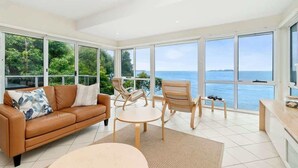 A modern living room with a leather sofa, wooden chairs, a round table, and large windows offering a view of the sea. - Tollgate Beach House (Denhams Beach)