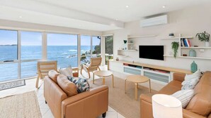 A modern living room with a large window overlooking the sea, a flat-screen TV, and a minimalist decor. - Tollgate Beach House (Denhams Beach)