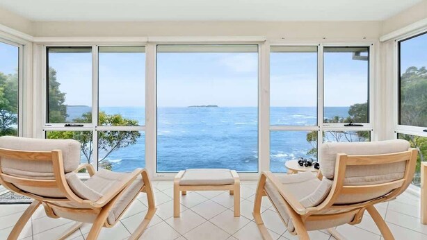A room with large windows, two wooden chairs, a small table, and a view of the sea. - Tollgate Beach House (Denhams Beach)